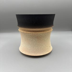 Puede incluir: Un jarrón de cerámica hecho a mano con un diseño único. La sección superior es negra, mientras que la parte inferior es de color crema con una apariencia agrietada y texturizada. El jarrón tiene una parte superior acampanada y una forma redondeada de reloj de arena.