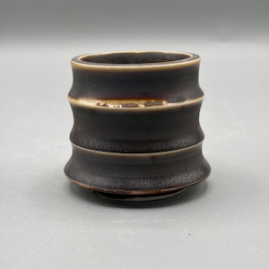 May include: A small, cylindrical ceramic cup featuring a dark brown, matte-textured finish with two horizontal ridges. The rim and the ridges exhibit a lighter, glossy tan glaze, creating a distinct contrast against the dark body of the vessel.