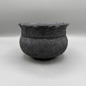 May include: A dark gray, round bowl with a textured, cratered surface and a scalloped, uneven rim. The exterior features a mottled, porous appearance with varying shades of charcoal and slate gray, creating a rugged, organic aesthetic.
