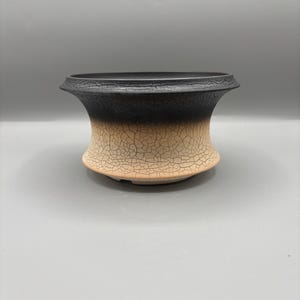 Puede incluir: Una maceta de cerámica hecha a mano con un esmalte craquelado único. La parte superior es negra, que pasa a una base beige. La maceta tiene un borde acampanado y una forma bulbosa, adecuada para plantas o uso decorativo.