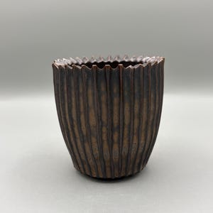 May include: A small, brown ceramic vase with a textured, ribbed exterior. The rim of the vase has a jagged, scalloped edge. The vase is approximately 4 inches tall and 4 inches wide, with a dark brown color.