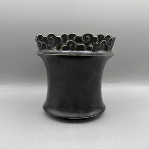 May include: A dark charcoal gray ceramic vessel with a flared rim featuring a raised, sculpted cloud pattern around the top edge. The body of the container has a smooth, matte finish that transitions into a slightly wider base.