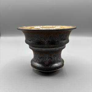 May include: A ceramic bowl with a flared rim, featuring a dark, textured exterior glaze in shades of black and deep brown. The interior of the bowl displays a smooth, contrasting light tan or beige glaze, creating a distinct visual separation.