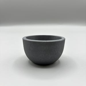 May include: A dark gray, semi-spherical bowl with a textured interior. The bowl has a rounded base and a slightly flared rim. The interior features horizontal ridges. The bowl is made of a solid, matte material.