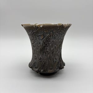 May include: A ceramic vessel with a flared rim and a dark, textured exterior featuring a mottled brown and black glaze. The surface has a rugged, organic appearance with deep grooves and raised ridges that create a complex, uneven tactile finish.