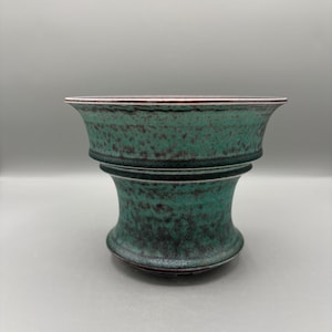 May include: A teal ceramic vase with a speckled texture. The vase has a wide, flared top and a rounded base, connected by a narrower midsection. The rim is a light pink colour.