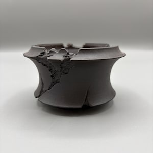 May include: A dark brown, handmade ceramic bowl with a unique, cracked design. The bowl has a wide, hourglass shape with a broken rim, adding a rustic and artistic touch. The dark color and texture suggest a natural, earthy aesthetic.