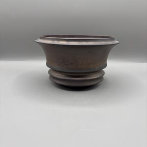 Puede incluir: Una maceta de cerámica marrón con un borde ancho y acanalado y una superficie texturizada. La maceta tiene una forma bulbosa con una serie de bandas redondeadas apiladas en la base. El color es marrón oscuro y terroso con sutiles variaciones de tono.