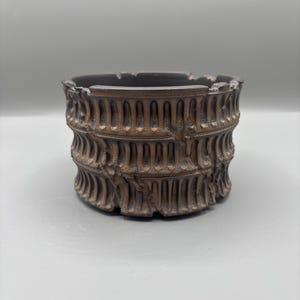 May include: A bronze-colored ashtray with a detailed, textured design. The ashtray has a cylindrical shape with a wide opening and a decorative pattern around the sides. The design appears to be inspired by classical architecture.
