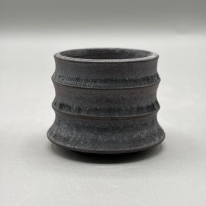 Puede incluir: Un pequeño recipiente cilíndrico de cerámica gris carbón oscuro con un exterior texturizado que presenta tres crestas horizontales distintas. La superficie tiene un acabado mate ligeramente rugoso con estrías verticales visibles en el cuerpo.