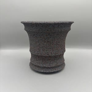 May include: A dark gray ceramic vase with a textured surface and a flared top. The vase has a cylindrical shape with a series of horizontal ridges near the base. The texture includes small flecks of a reddish-brown color.