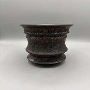 Puede incluir: Un cuenco de cerámica de color marrón oscuro con un borde ancho y acampanado y un cuerpo en forma de reloj de arena con nervaduras. La superficie presenta un esmalte texturizado y moteado con tonos bronce y negro metálicos.