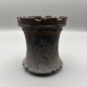 May include: A dark brown ceramic tower-shaped vase with a textured surface. The vase has a flared base and a crenelated rim, resembling a castle turret. The color gradient transitions from dark brown to a lighter shade near the base.