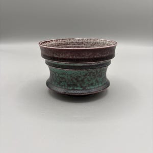May include: A small ceramic bowl with a flared rim, featuring a dark purple-brown upper section and a textured, mottled teal-green lower body. The interior rim shows horizontal ridges, and the overall finish has a matte, speckled appearance.