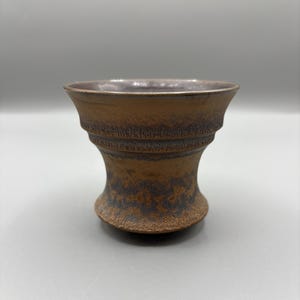 May include: A brown ceramic bowl with a textured surface and a flared rim. The bowl has a wide base and a narrower, decorative midsection. The interior of the bowl has a lighter color than the exterior.