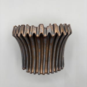 May include: A decorative, bronze-colored bowl with a fluted design. The bowl has a wide, flared shape with vertical ridges that create a textured surface. The rim has a scalloped edge, adding to its ornate appearance.