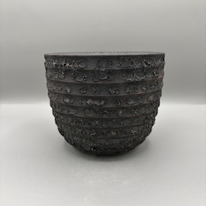 May include: A dark gray, textured ceramic bowl with a rounded shape. The bowl features horizontal bands with a rough, pitted surface. The interior is a darker shade of gray. The bowl is likely handmade and suitable for decorative or functional use.