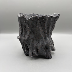 May include: A dark charcoal gray planter with a rough, uneven, and textured exterior surface that mimics the appearance of tree bark. The vessel features a wide, open top and a sturdy base with small, integrated feet for stability.