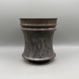 May include: A cylindrical ceramic vessel with a flared rim and base, featuring a dark, mottled metallic glaze with copper and charcoal gray tones. The surface has a textured, uneven finish with visible color variations throughout the body.