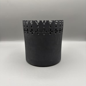 Może przedstawiać: Cylindryczny czarny ceramiczny pojemnik o teksturowanej powierzchni. Górna krawędź ma dekoracyjny, karbowany wzór z małymi okrągłymi i krzyżowymi wycięciami. Nadaje się do przechowywania małych przedmiotów lub jako element dekoracyjny.