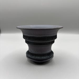 May include: A dark gray decorative vase with a flared rim and a detailed, textured design. The vase has a wide opening and a rounded base, with horizontal bands of intricate patterns. The overall aesthetic is classic and elegant.