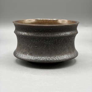 May include: A dark, metallic-toned ceramic bowl with a distinct hourglass-shaped silhouette. The exterior features a textured, speckled finish in shades of charcoal and bronze, while the interior has a smooth, reflective metallic copper-colored glaze.