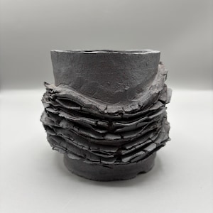 May include: A dark gray, handcrafted ceramic planter with a unique, textured design. The planter features a cylindrical shape with a layered, organic texture around the base, creating a rustic and modern aesthetic. Ideal for small plants or succulents.