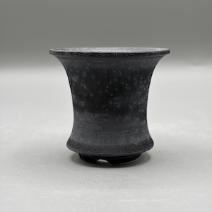 May include: A small, flared ceramic pot with a matte black finish and a textured, speckled surface. The vessel features a wide, open rim that tapers inward toward a narrow base, which includes small cutouts along the bottom edge for drainage.