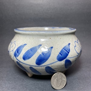 Puede incluir: Una pequeña maceta de cerámica gris con diseños florales azules y tres patas. La maceta es del tamaño de una moneda de 1 euro.