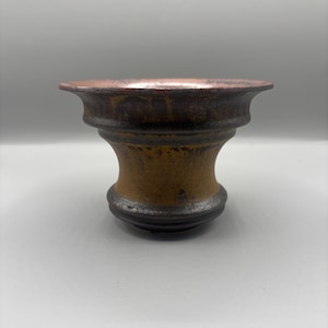 May include: A handcrafted ceramic vase with a flared rim and a bulbous body. The vase features a gradient of colors, transitioning from a dark brown base to a golden-brown midsection and a reddish-brown rim. The textured surface adds to its rustic charm.