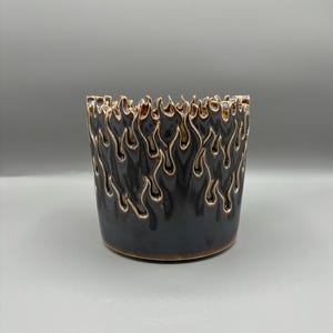 May include: A cylindrical ceramic planter featuring a glossy dark brown glaze with a raised, textured flame pattern around the top rim. The flames are outlined in a lighter tan color, creating a distinct contrast against the dark body of the vessel.