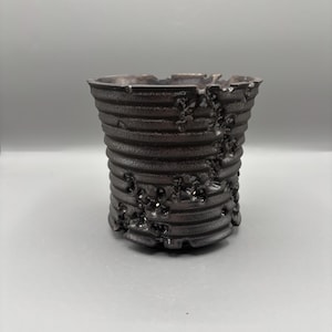 Puede incluir: Una maceta cilíndrica de color gris oscuro con un exterior texturizado y acanalado. El borde superior es irregular y la superficie tiene pequeños agujeros irregulares. La maceta está diseñada para uso en interiores o exteriores.