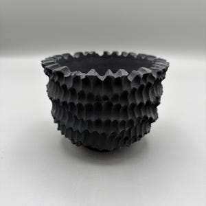 May include: A small, matte black bowl featuring a deeply textured, scalloped exterior surface with repeating vertical ridges. The bowl has a wide, open circular top with a jagged, uneven rim and a rounded base, presenting a uniform dark colour.