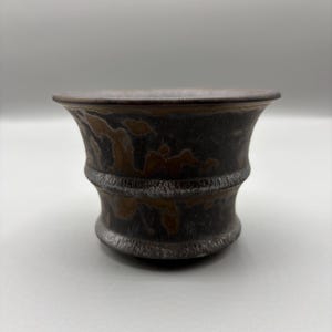 Pode incluir: Uma pequena tigela de cerâmica marrom escuro com uma borda alargada e uma aparência texturizada e envelhecida. A tigela tem uma base cilíndrica com um design canelado. O interior é um tom mais escuro de marrom, e o exterior tem um acabamento rústico manchado.