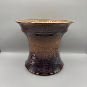 May include: A handcrafted ceramic vase with a wide, flared rim and a bulbous body. The vase features a gradient of brown and burgundy hues, with a glossy finish. The base has a subtle, golden rim.