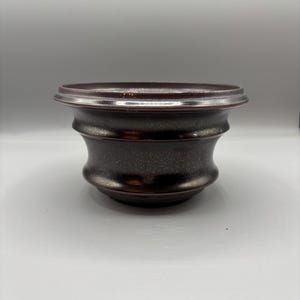 May include: A dark brown ceramic bowl with a wide rim and a textured surface. The bowl has a rounded shape with a slightly flared base. The rim has a silver-colored band. The bowl is likely used for serving or display.
