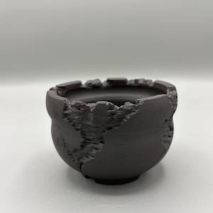 May include: A dark gray, small, round ceramic planter with a textured, distressed exterior. The rim has a jagged, uneven edge. The planter has a small, circular base and a smooth interior. The overall design gives the appearance of a weathered, aged aesthetic.
