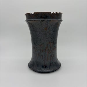 May include: A dark gray ceramic vase with a textured surface and a flared base. The vase has a unique, jagged rim and a metallic sheen, adding a touch of rustic elegance. The vase is approximately 8 inches tall.