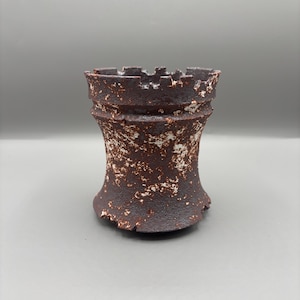 May include: A dark brown ceramic rook chess piece with a distressed, textured finish. The chess piece has a cylindrical body with a flared base and a crenellated top. White accents are scattered across the surface.