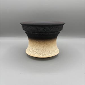 May include: A ceramic vase with a unique, textured design. The top is dark brown, transitioning to black, and then to a beige base. The surface has a cracked, aged appearance, adding to its artistic appeal.