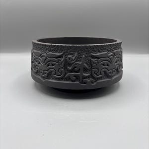 Puede incluir: Un cuenco decorativo redondo de color gris oscuro con un intrincado patrón repetitivo tallado alrededor del exterior. El cuenco tiene una abertura ancha y una base ligeramente curvada. El diseño parece estar inspirado en el arte antiguo.