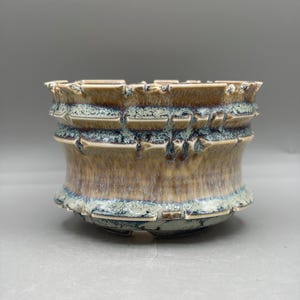 May include: A ceramic planter with a wide, cylindrical shape and a textured, notched rim. The surface features a reactive glaze with blended tones of tan, beige, and deep blue, creating a mottled, layered appearance across the exterior.
