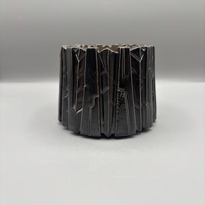 Puede incluir: Un recipiente decorativo cilíndrico de color marrón oscuro con un diseño geométrico texturizado. El recipiente tiene un exterior rugoso y angular, con una serie de formas verticales e irregulares. El interior es liso y parece ser de un tono más claro de marrón. El recipiente es probablemente de cerámica o metal.