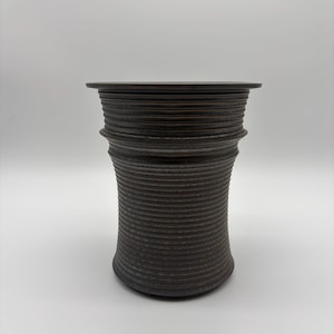May include: A dark brown cylindrical vessel with a textured, ribbed surface featuring horizontal grooves throughout. The object has a wide, flat rim at the top and a slightly tapered body that widens toward the base, showcasing a matte finish.
