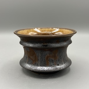 May include: A handcrafted ceramic bowl with a unique design. The bowl features a dark brown exterior with a metallic silver and brown interior. The rim is a lighter shade of brown. The bowl has a wide, rounded shape.