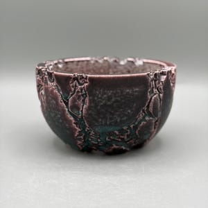 May include: A small, round ceramic bowl featuring a dark, textured exterior with deep fissures and a metallic, iridescent finish in shades of plum, copper, and dark green. The rim is uneven and rugged, showcasing a complex, cracked glaze pattern.