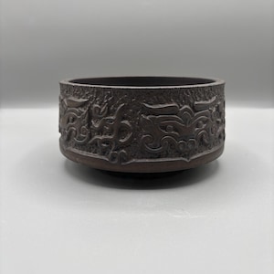 May include: A dark brown, round, decorative bowl with an embossed design. The bowl's exterior features a repeating pattern of stylized dragon-like figures. The bowl has a low profile and sits on a slightly raised base.