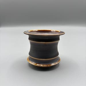 May include: A dark brown ceramic vessel with a wide, flat rim and a rounded base. The body of the vessel has a cylindrical shape with a slight hourglass curve. The rim and base have a lighter brown glaze, creating a contrast with the darker body.