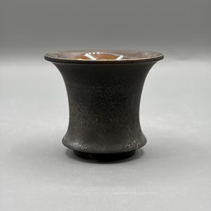 May include: A small, flared ceramic cup with a dark, textured exterior finish that transitions to a smooth, glossy brown glaze on the interior rim. The vessel features a wide, open top that tapers inward toward a narrow, circular base.