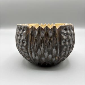 May include: A textured, dark brown bowl with a gold interior. The bowl has a unique, organic pattern on its exterior and a jagged edge around the rim. The bowl is round and sits on a white surface.